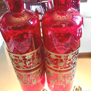 May include: Two red glass bottles with gold metal filigree designs around the middle. The bottles are sitting on a gold and white patterned surface.