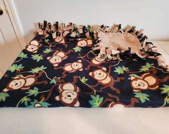 May include: A fleece tie blanket featuring a dark blue background with a repeating pattern of brown and cream-colored monkeys and green leaves. The edges of the blanket are finished with knotted fringe, alternating between dark blue and solid tan fabric.