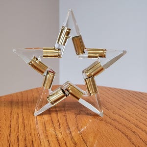 May include: A clear acrylic star sculpture with brass bullet casings embedded within the points. The star is displayed on a wooden surface, showcasing a unique blend of materials and textures. The sculpture is a decorative item.
