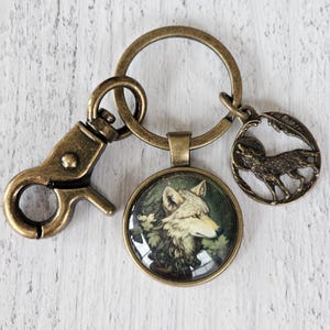 May include: A bronze-toned keychain featuring a circular pendant with a detailed illustration of a wolf's head, a lobster clasp, and a smaller circular charm depicting a wolf silhouette against a moon, all finished in an antique bronze metal.