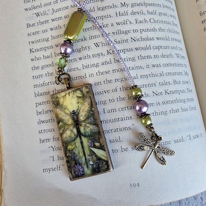 May include: A bookmark featuring a rectangular pendant with a dragonfly illustration, accented by a purple cord and beads in shades of green and lavender. A small metal dragonfly charm hangs from the beaded strand, which is draped over an open book.