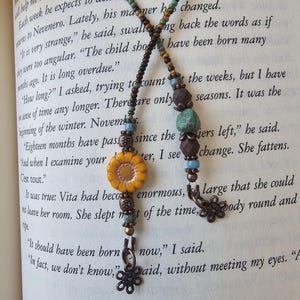 May include: A beaded bookmark featuring two strands of small glass beads in shades of green, brown, and blue. One strand ends with a yellow ceramic sunflower bead and a copper flower charm, while the other features a turquoise stone and copper flower.