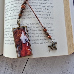 May include: A bookmark featuring a rectangular pendant with a horse illustration, attached to a beaded cord with copper-toned beads and a small bronze-colored horse charm. The cord is made of small brown beads and rests on an open book page.
