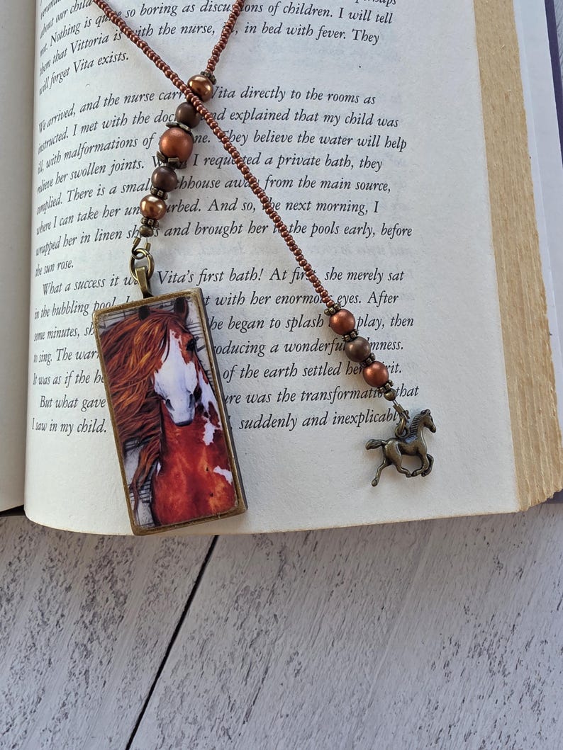 May include: A bookmark featuring a rectangular pendant with a horse illustration, attached to a beaded cord with copper-toned beads and a small bronze-colored horse charm. The cord is made of small brown beads and rests on an open book page.