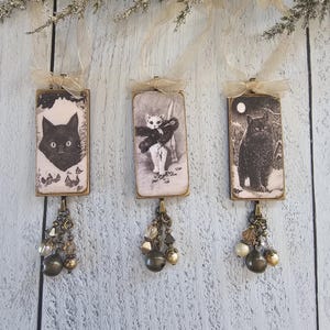 May include: Three rectangular hanging ornaments featuring black and white cat illustrations. Each ornament has a gold-toned metal frame, a sheer beige ribbon loop at the top, and a dangling charm cluster of metallic beads and crystals at the bottom.