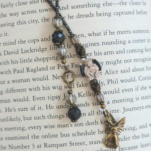 May include: A beaded necklace with a dark chain and various beads in black, silver, and brown. The pendant features a small, bronze-colored winged figure. The necklace is displayed on a book page.