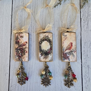 May include: Three rectangular hanging ornaments featuring vintage-style illustrations of birds and a wreath. Each piece has a gold ribbon loop, a bronze-toned metal frame, and a dangling charm cluster with colorful beads and a metal snowflake.