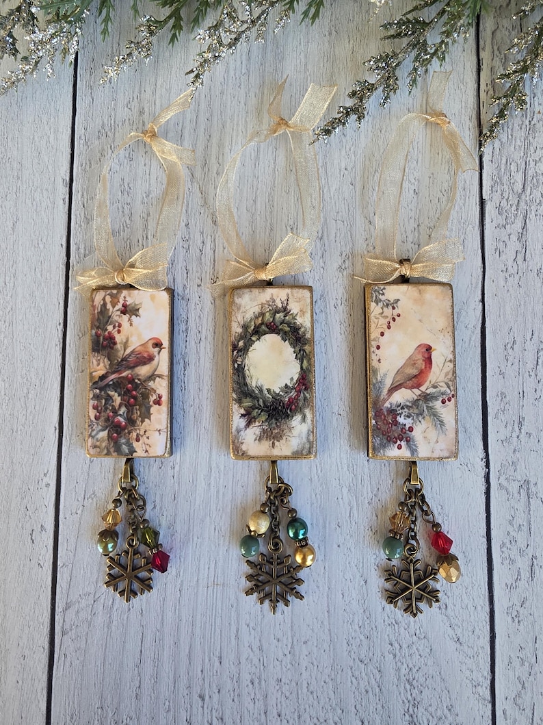 May include: Three rectangular hanging ornaments featuring vintage-style illustrations of birds and a wreath. Each piece has a gold ribbon loop, a bronze-toned metal frame, and a dangling charm cluster with colorful beads and a metal snowflake.