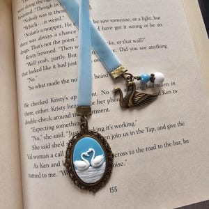 May include: A light blue velvet ribbon bookmark featuring an oval cameo pendant with two white swans on a blue background, framed in antique bronze. A secondary bronze swan charm and small blue and white beads are attached to the ribbon end.