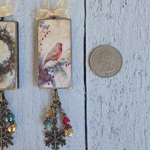 May include: Two rectangular hanging ornaments featuring a vintage-style illustration of a red bird perched on a pine branch with berries. Each ornament has a gold ribbon loop, a bronze-toned chain, colorful beads, and a metal snowflake charm.
