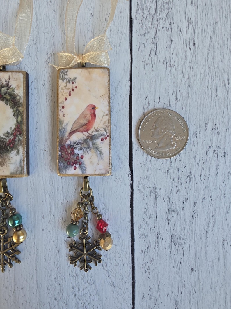 May include: Two rectangular hanging ornaments featuring a vintage-style illustration of a red bird perched on a pine branch with berries. Each ornament has a gold ribbon loop, a bronze-toned chain, colorful beads, and a metal snowflake charm.