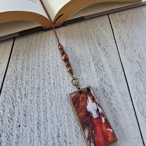 May include: A rectangular bookmark featuring a printed image of a chestnut horse with a white blaze on its face. The pendant is attached to a brown cord adorned with three metallic copper-colored beads, resting inside an open hardcover book.