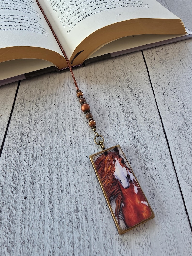 May include: A rectangular bookmark featuring a printed image of a chestnut horse with a white blaze on its face. The pendant is attached to a brown cord adorned with three metallic copper-colored beads, resting inside an open hardcover book.