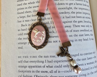 May include: A pink velvet ribbon bookmark featuring an oval bronze-toned cameo pendant with a white hummingbird and floral relief. The bookmark includes a bronze hummingbird charm and a dangling pearl-like bead attached to the ribbon end.