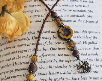 May include: A beaded bookmark featuring a long strand of small brown beads, accented with larger yellow and brown spherical beads, a central yellow flower-shaped bead, and two bronze-toned metal bee charms at each end of the beaded cord.