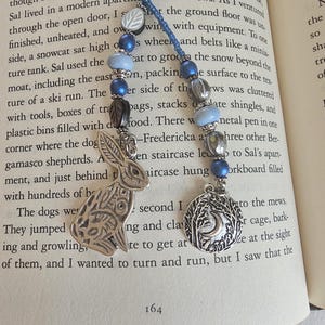 May include: A beaded bookmark featuring a blue cord with two strands of blue, silver, and white beads. One strand ends in a silver-toned metal rabbit charm, while the other strand features a round silver-toned charm depicting a crescent moon and trees.