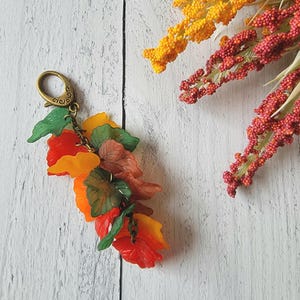 May include: A keychain featuring a cluster of translucent acrylic leaves in shades of orange, yellow, green, and brown, attached to a bronze-toned metal lobster clasp. The leaves are arranged in a layered, cascading design with visible veins.