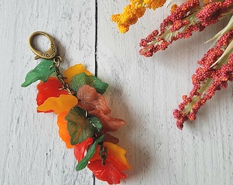 May include: A keychain featuring a cluster of translucent acrylic leaves in shades of orange, yellow, green, and brown, attached to a bronze-toned metal lobster clasp. The leaves are arranged in a layered, cascading design with visible veins.