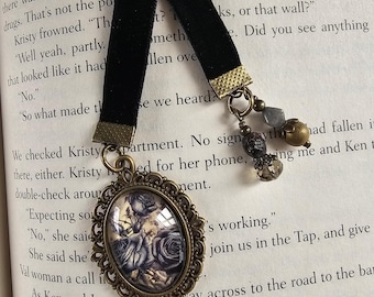 May include: A black velvet ribbon bookmark featuring an oval antique bronze pendant with a floral rose print under a glass cabochon. The bookmark includes a dangling charm cluster with small grey, brown, and clear beads attached to the ribbon end.