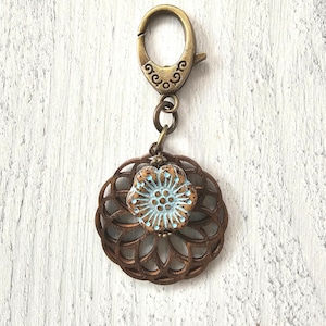 May include: A bronze-toned metal lobster clasp attached to a circular, open-work wooden pendant. Centered on the wooden piece is a small, round, speckled ceramic bead featuring a light blue floral pattern with a raised, textured surface.