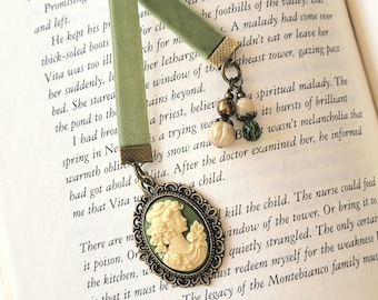 May include: A sage green velvet ribbon bookmark featuring an oval antique bronze frame with a cream-colored cameo relief of a profile. The bookmark is accented with a dangling charm cluster of two small beads in cream and green tones.