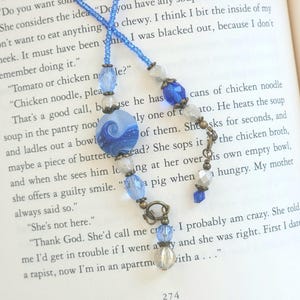 May include: A blue beaded bookmark with a wave design on a round glass bead. The bookmark features various blue and clear beads, with a bronze-colored metal clasp. The bookmark is resting on the pages of a book.