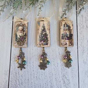 May include: Three rectangular hanging ornaments featuring vintage-style Christmas imagery of Santa Claus, a decorated tree, and stacked gift boxes. Each piece is topped with a sheer gold ribbon and finished with a dangling bronze snowflake charm.