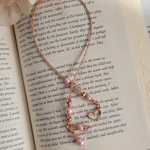 May include: A rose gold chain bookmark featuring a delicate beaded design with soft pink pearls, faceted clear beads, and a small open heart charm. The bookmark is draped over an open book page with printed text, showcasing a refined and elegant aesthetic.