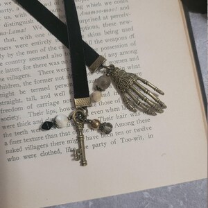May include: A black velvet ribbon bookmark featuring two dangling charms: a metallic skeleton hand and a vintage-style skeleton key. The charms are accented with small, multi-colored beads in shades of black, white, and bronze, attached with metal findings.