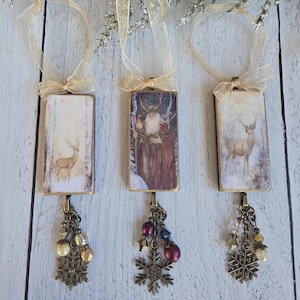 May include: Three rectangular hanging ornaments with gold-toned edges and sheer gold ribbons. Ornament 1 features a deer in a snowy forest. Ornament 2 depicts a bearded figure in a red cloak. Ornament 3 shows a deer. Each has a bronze snowflake charm.