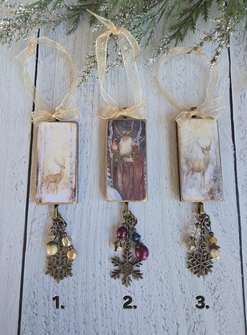 May include: Three rectangular hanging ornaments with gold-toned edges and sheer gold ribbons. Ornament 1 features a deer in a snowy forest. Ornament 2 depicts a bearded figure in a red cloak. Ornament 3 shows a deer. Each has a bronze snowflake charm.