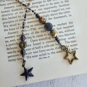 May include: A close-up of a necklace with a variety of beads, including black, gold, and brown. The necklace has two star-shaped charms, one black and one gold. The necklace is lying on a book with text about the Antarctic circle. The text on the book reads "January 12th. Our passage to the south again looked doubtful, as nothing was to be seen in the direction of the blue mountains of ragged ice, one precipice of which arose frowningly above the other."