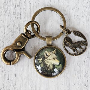 May include: Antique brass keychain featuring a round pendant with a wolf portrait, a wolf silhouette charm, and a lobster clasp. The wolf pendant has a glass dome and a forest background. The keychain is a rustic accessory.