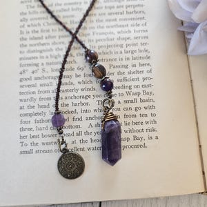 May include: A necklace featuring a dark brown braided cord, a series of round beads in shades of purple and brown, a small round antique bronze-toned metal charm with an intricate pattern, and a purple crystal point wrapped in bronze wire.