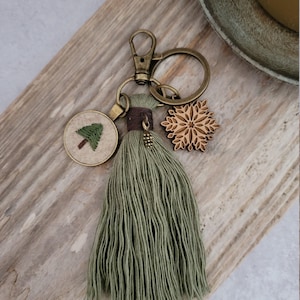 May include: A keychain with a green tassel, a round charm with an embroidered tree, and a wooden snowflake charm. The keychain has a bronze-colored clasp and ring. A small bronze-colored pinecone charm is attached to the tassel.