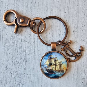 May include: A bronze-colored keychain with a round glass charm featuring a vintage sailing ship. The charm is attached to a split ring with a lobster clasp and a small anchor charm.
