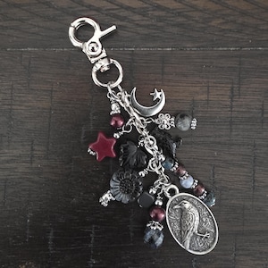 May include: A silver-toned metal keychain featuring a lobster clasp and multiple dangling charms, including a crescent moon, a star, a black flower, a dark red star, and an oval pendant embossed with a detailed raven illustration on a scrollwork background.
