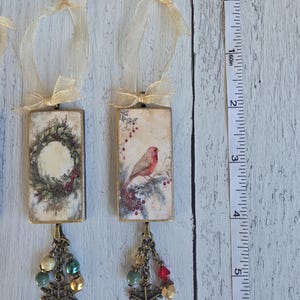 May include: Two rectangular hanging ornaments with gold-toned metal frames and sheer beige ribbons. One features a wreath, the other a bird on a branch. Both have dangling bronze-toned snowflake charms and beads. A measuring tape shows a 6-inch length.