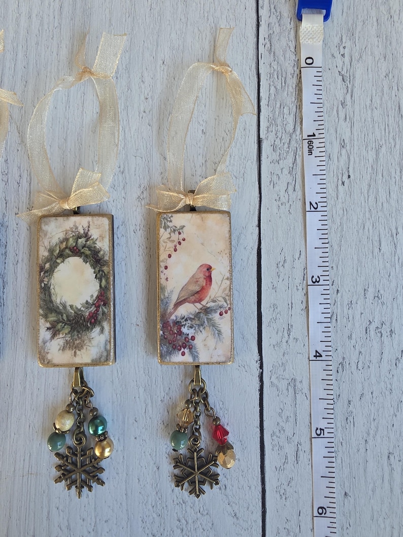 May include: Two rectangular hanging ornaments with gold-toned metal frames and sheer beige ribbons. One features a wreath, the other a bird on a branch. Both have dangling bronze-toned snowflake charms and beads. A measuring tape shows a 6-inch length.