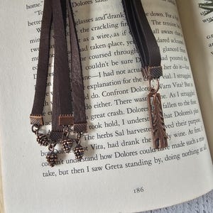 May include: Brown leather bookmarks with bronze-colored pinecone and feather charms. The bookmarks are displayed on the open pages of a book. The page number 186 is visible.
