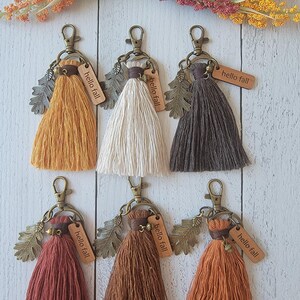 May include: Six macrame tassel keychains in mustard, cream, dark gray, rust, brown, and orange. Each features a bronze-toned lobster clasp, a metal oak leaf charm, a small acorn charm, and a wooden rectangular tag engraved with the text hello fall.