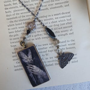 May include: A necklace featuring a rectangular pendant with a dark image of a hand holding a moth, accented by a small metal moth charm. The chain is composed of small grey, black, and clear beads, with a larger black oval bead and a clear faceted bead.