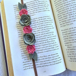 May include: A handmade fabric bookmark with felt roses in pink and gray, accented with green leaves. The bookmark is attached to the pages of a book, adding a decorative touch to reading.