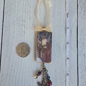 May include: A rectangular hanging ornament featuring an illustration of a bearded figure with antlers in a snowy forest. It has a gold ribbon loop at the top and a bronze-toned charm cluster with beads and a snowflake at the bottom, measuring 4 inches.