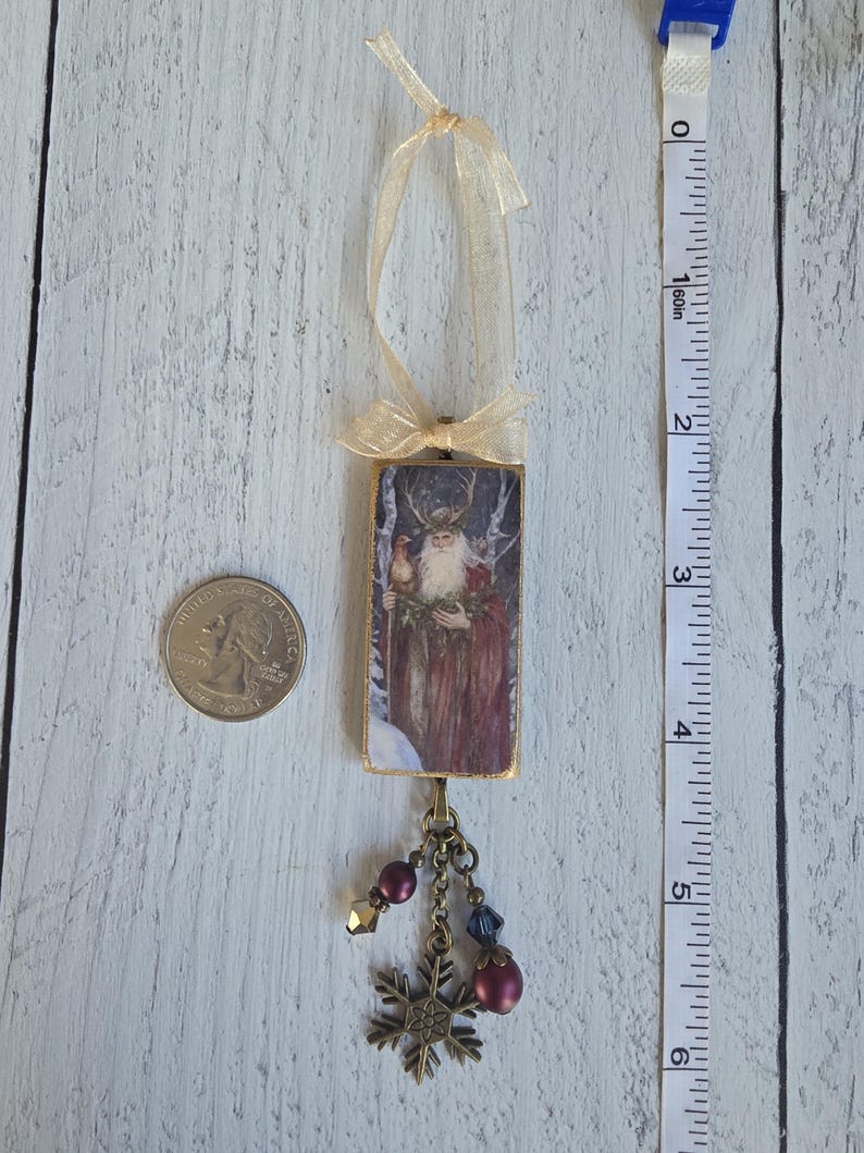 May include: A rectangular hanging ornament featuring an illustration of a bearded figure with antlers in a snowy forest. It has a gold ribbon loop at the top and a bronze-toned charm cluster with beads and a snowflake at the bottom, measuring 4 inches.