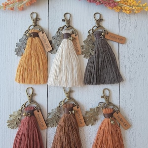 May include: Six fall-themed keychains with tassels in various colors: mustard yellow, cream, dark brown, rust, medium brown, and orange. Each keychain has a metal leaf charm, a small wooden tag that says "hello fall", and a clasp.