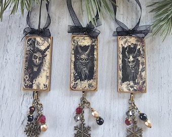 May include: Three rectangular hanging ornaments featuring a black and white illustration of a horned creature. Each piece has a black ribbon loop, a bronze-toned metal chain, assorted red, black, and gold beads, and a bronze snowflake charm.