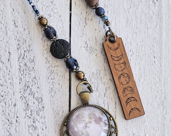 Op de afbeelding: Een ketting met een ronde hanger met een maanafbeelding in een bronskleurig sterrenframe, bevestigd aan een kralensnoer met blauwe, zwarte en bruine kralen. Een rechthoekige houten bedel met maanfasen hangt aan het kralensnoer.
