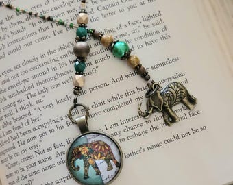 May include: A bookmark featuring a beaded chain with green, tan, and metallic beads. One end has a round pendant with a colourful elephant illustration, and the other end features a bronze-toned metal elephant charm with intricate etched details.