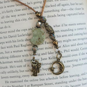May include: A beaded necklace featuring a bronze chain, a pale green crescent moon stone, and bronze charms including two owls and a crescent moon with a star. The necklace is adorned with small metallic beads and faceted glass accents.